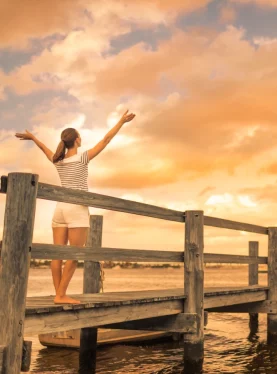 A woman with open arms on a pier by the water, symbolizing freedom and self-discovery through Thrive Therapy in Weybridge and online.